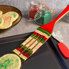 A multicolored kitchen tool from the Totally Bamboo Baltique® North Pole Collection, the 13" Spurtle, rests on a baking sheet next to sugar cookies sprinkled with festive Christmas coloring. In the background, glass jars contain vibrant red and green sprinkles.