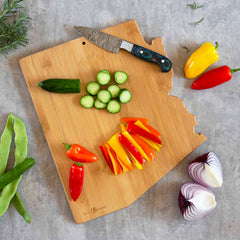 The Arizona Cutting Board with Artwork by Fish Kiss™ from Totally Bamboo showcases sliced cucumbers and vibrant red and yellow pepper strips, accompanied by a knife resting nearby. Whole peppers, cucumbers, and halved red onions are scattered around, while fresh herbs peek from the corners to add a touch of greenery to this culinary canvas.