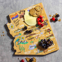 The Arizona Cutting Board with Artwork by Fish Kiss™ from Totally Bamboo showcases colorful illustrations of landmarks and towns in Arizona. The bamboo surface beautifully displays cheese, crackers, cherries, blackberries, and grapes against a gray background.