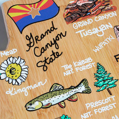 The Arizona Cutting Board with Artwork by Fish Kiss™, from the brand Totally Bamboo, showcases a decorative state-shaped design featuring illustrations and labels of Arizona attractions such as the Grand Canyon, Lake Mead, Kingman, Tusayan, Prescott National Forest, and Kaibab National Forest. The Arizona state flag is prominently displayed at the top.