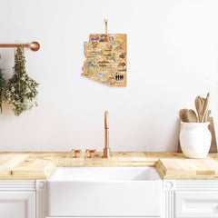 A modern kitchen features a wooden countertop with a large white farmhouse sink, complemented by an Arizona Cutting Board with Artwork by Fish Kiss™ from Totally Bamboo. Above, an Arizona map art piece adorns the white wall. Copper faucets, herbs, and kitchen utensils are neatly organized around the sink to complete the stylish setup.