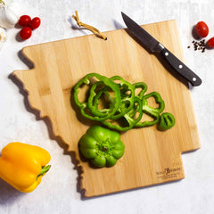 The Arkansas Cutting Board with Artwork by Fish Kiss™, crafted by Totally Bamboo, displays sliced green bell pepper with a whole one beside it, while a knife rests nearby. Cherry tomatoes, garlic, and a yellow bell pepper provide colorful accents to the marble surface. The board is adorned with vibrant graphics thanks to its food-safe UV printing.