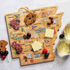 The Arkansas Cutting Board with Artwork by Fish Kiss™, crafted in the shape of the state, showcases local landmarks and cities. This wooden masterpiece from Totally Bamboo is vibrantly decorated with cheese slices, crackers, nuts, sausage, cherries, and two wine glasses for an elegant charcuterie display.