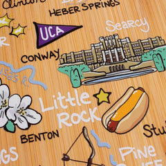 The Arkansas Cutting Board with Artwork by Fish Kiss™ from Totally Bamboo showcases an illustrated map adorned with Arkansas landmarks and icons such as a hot dog, "Little Rock," and a university building. It features text highlighting cities like Conway, Heber Springs, and Searcy. The bamboo-like background enhances the charm of this piece, which is crafted using Food-Safe UV Printing techniques.