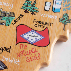 The Arkansas Cutting Board with Artwork by Fish Kiss™, crafted by Totally Bamboo, is made using food-safe UV printing and showcases illustrations of local landmarks, the state flag, and cities such as Searcy, Forrest City, and DeWitt. The phrase "The Natural State" is prominently featured alongside the flag.