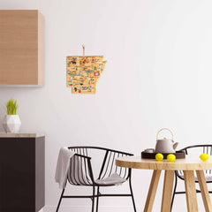 A modern kitchen features a wooden cabinet, a black chair with a gray blanket, and a round table with a teapot and yellow cups. A decorative map of Arkansas hangs on the white wall. On the countertop, you'll find an Arkansas Cutting Board with Artwork by Fish Kiss™ from Totally Bamboo alongside a potted plant and hexagonal vase.