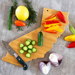 A California Cutting Board with Artwork by Fish Kiss™, crafted from a durable bamboo cutting surface by Totally Bamboo, holds sliced bell peppers and cucumbers. Surrounding it are whole cucumbers, a lemon, rosemary, a knife, a halved onion, and various peppers on a gray surface.