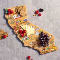 The California Cutting Board with Artwork by Fish Kiss™ from Totally Bamboo features an elegant bamboo surface shaped like the state of California. It beautifully displays vibrant artwork depicting cities and landmarks, serving as a perfect base for arranging cheese slices, grapes, cookies, and berries against a gray marble backdrop.