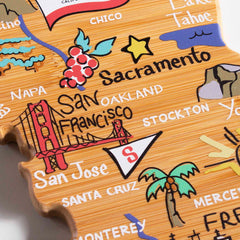 Introducing the California Cutting Board with Artwork by Fish Kiss™, a unique bamboo cutting surface crafted in the shape of the state. This Totally Bamboo product showcases charming illustrations of cities including San Francisco, Sacramento, and San Jose, as well as doodles like the Golden Gate Bridge, grapes, and a surfing palm tree for added flair.