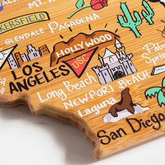 The California Cutting Board with Artwork by Fish Kiss™ from Totally Bamboo is a wooden cutout map of California, similar to state-shaped cutting boards or bamboo surfaces. It features illustrated cities such as Los Angeles and San Diego, along with icons like the Hollywood sign and a theme park castle nestled among palm trees and cacti.