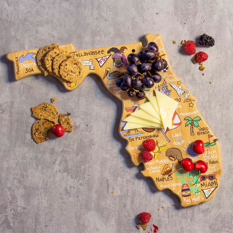 The Florida Cutting Board with Artwork by Fish Kiss™ from Totally Bamboo beautifully displays cities and landmarks with vibrant designs. Its bamboo surface is perfect for holding grapes, cheese slices, cookies, and berries, while scattered berries and cookies adorn the gray surface below.