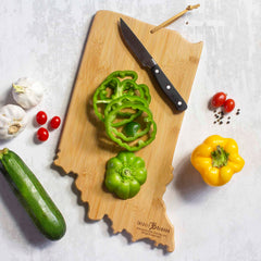 A bamboo cutting board, branded as the Indiana Cutting Board with Artwork by Fish Kiss™ from Totally Bamboo, is shaped like the Indiana state map and displays sliced green bell peppers alongside a knife. Surrounding the board are a whole green pepper, a yellow pepper, zucchini, garlic, cherry tomatoes, and peppercorns on a light surface.