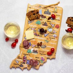 The Indiana Cutting Board with Artwork by Fish Kiss™ from Totally Bamboo, featuring an intricate design of city names and illustrations, holds cheese, salami, cherry tomatoes, and crackers. Next to it on a white surface are two glasses of white wine.