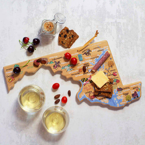 The Maryland-shaped cutting board from Totally Bamboo, featuring vibrant Fish Kiss™ artwork, is beautifully crafted and adorned with cheese, salami, tomatoes, and crackers. Nearby are cherries and a jar of toothpicks alongside two glasses of white wine on a textured surface.