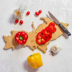 On the Michigan Upper Peninsula Cutting Board with Artwork by Fish Kiss™ by Totally Bamboo, sliced red bell peppers rest in a design reminiscent of a geographical map. Nearby, a knife is surrounded by cherry tomatoes and a yellow bell pepper. A jar of toothpicks and a bulb of garlic complete the scene on the textured surface.