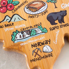 Discover the Michigan Upper Peninsula Cutting Board with Artwork by Fish Kiss™, a creation by Totally Bamboo. This illustrated wooden masterpiece showcases landmarks and symbols such as a pie, bear, and mountains. The artwork includes labels for "Iron River," "Crystal Falls," "Norway," and "Menominee," while trees and tools highlight local attractions on this elegantly crafted bamboo surface shaped like state-shaped cutting boards.