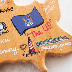 The Michigan Upper Peninsula Cutting Board with Artwork by Fish Kiss™, crafted by Totally Bamboo, features a decorative design that highlights the Upper Peninsula with colorful illustrations, such as the state flag, a lighthouse, and Mackinac Island. Handwritten labels accompany each feature to provide a playful and informative touch.