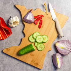 A Minnesota-shaped cutting board by Totally Bamboo, featuring artwork from Fish Kiss™, showcases sliced cucumber, a halved onion, a bulb of garlic, and a red pepper. A knife is placed next to it on the gray surface, where additional sliced vegetables and garlic are scattered. This piece is perfect for those who appreciate kitchen art.