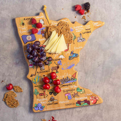 The Minnesota Cutting Board with Artwork by Fish Kiss™ from Totally Bamboo, featuring a bamboo cutting surface shaped like Minnesota, is adorned with a variety of cheeses, crackers, cherries, and grapes. Colorful drawings and labels of the state's cities and landmarks enhance the board. Additional berries and crackers are scattered around.