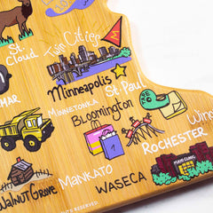 The Minnesota Cutting Board with Artwork by Fish Kiss™ from Totally Bamboo features vibrant illustrations of cities like Minneapolis, St. Paul, Rochester, and Waseca. Designed by Fish Kiss Artwork, it showcases buildings and trees alongside city names on a functional state-shaped surface that's both practical and decorative.