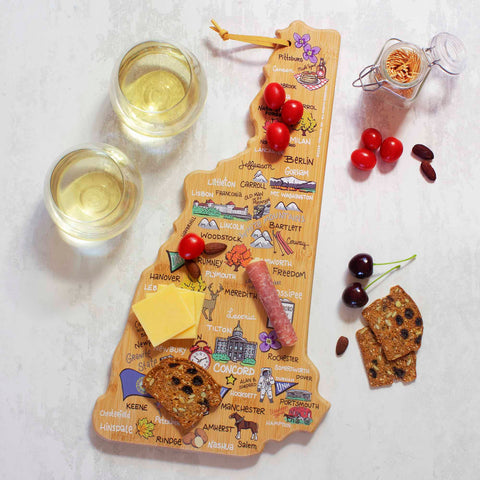 The New Hampshire Cutting Board by Totally Bamboo, featuring Fish Kiss™ artwork and made from bamboo cutting surface materials, showcases a delightful spread of crackers, cheese, salami slices, cherries, and tomatoes. Adjacent on a light surface sit two glasses of white wine and a jar of crackers.