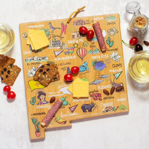 A New Mexico State Shaped Cutting and Serving Board from Totally Bamboo, featuring vibrant artwork by Fish Kiss™, is elegantly topped with an array of cheese, crackers, cherry tomatoes, and salami. Complemented by two glasses of white wine, it's an ideal setup for a serene evening.