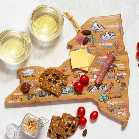 The New York Cutting Board with Artwork by Fish Kiss™ from Totally Bamboo features a wooden state-shaped design, showcasing cheese, a salami stick, cherry tomatoes, and crackers. Accompanying the spread are two glasses of white wine and a jar of nuts. This setup is perfect for highlighting iconic locations while indulging in delicious snacks.