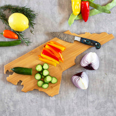 A North Carolina cutting board crafted by Totally Bamboo, featuring artwork by Fish Kiss™, elegantly showcases sliced cucumbers and bell peppers. A knife rests nearby, surrounded by whole vegetables, herbs, and a halved red onion on a grey surface—a perfect blend of form and function for fans of state-shaped cutting boards.