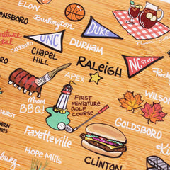 The North Carolina Cutting Board with Artwork by Fish Kiss™, crafted by Totally Bamboo, highlights playful Fish Kiss illustrations of iconic North Carolina symbols. It includes city names like RDU, Fayetteville, Chapel Hill, and Durham; sports emblems; musical instruments; BBQ; a burger; and seasonal leaves.