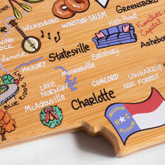 Introducing the North Carolina Cutting Board with Artwork by Fish Kiss™ from Totally Bamboo: a distinct wooden map adorned with vibrant illustrations and handwritten city names, including Charlotte and Statesville. This cutting board captures state pride perfectly, featuring the flag and local icons on a bamboo surface to showcase notable locations and features beautifully.