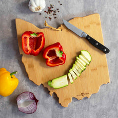 The Ohio Cutting Board with Artwork by Fish Kiss™ from Totally Bamboo features sliced zucchini and red bell pepper halves. A yellow bell pepper, half a red onion, garlic, and a small knife are placed nearby on the bamboo cutting surface. Peppercorns are scattered across the gray countertop.