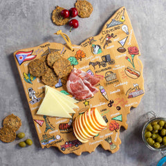 The Ohio Cutting Board with Artwork by Fish Kiss™ from Totally Bamboo, shaped like the state of Ohio and embellished with city names, serves as a stylish display for cheese slices, apples, cured meat, crackers, and a handful of cherries. Nearby on the gray surface sits a small jar of olives.