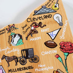 The Ohio Cutting Board with Artwork by Fish Kiss™ from Totally Bamboo beautifully highlights Ohio landmarks on a bamboo surface, showcasing elements such as a runner, football, horse and buggy, carnations, and city names including Cleveland, Akron, Canton, and Youngstown. It is vibrant and colorful with cultural symbols that echo the distinctive style of Fish Kiss Artwork.