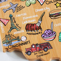 The Ohio Cutting Board with Artwork by Fish Kiss™, crafted by Totally Bamboo, features a vibrant depiction of colorful Ohio cities, including Dayton, Cincinnati, and Columbus. It highlights iconic markers such as an Eiffel Tower and a fire truck, complemented by whimsical elements like a plane and cheeseburger on this distinctive bamboo cutting surface.