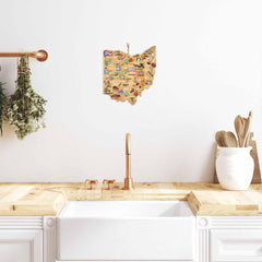 In a minimalist kitchen, a wooden countertop is paired with a white sink. A colorful Fish Kiss™ map of Ohio hangs on the white wall. Nearby, copper faucets and utensils gleam alongside a bamboo cutting board from Totally Bamboo, hanging herbs, and an Ohio-shaped cutting board featuring artwork by Fish Kiss™.