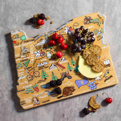 The Oregon Cutting Board with Artwork by Fish Kiss™, crafted by Totally Bamboo, showcases vivid depictions of Oregon's landmarks and cities. It is elegantly adorned with grapes, cherries, blackberries, cheese, and crackers on a stone surface.