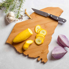 The South Carolina Cutting Board with Artwork by Fish Kiss™, from the brand Totally Bamboo, showcases hand-drawn designs while holding sliced yellow squash, a knife, and whole garlic cloves. Nearby, a sprig of rosemary and a half-cut red onion complement the sleek bamboo cutting surface against a pristine white background.