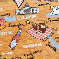 This hand-drawn artwork on the South Carolina Cutting Board with Artwork by Fish Kiss™ from Totally Bamboo features an illustrated map of Columbia. It includes a star, a building, and iced tea on a picnic cloth. Nearby are drawings such as hot sauce labeled "Orangeburg" and a swan, along with other locations like Camden, Sumter, Santee, and Lexington.