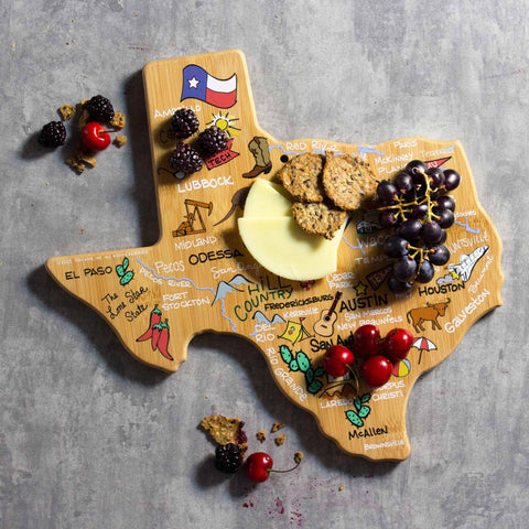 The Texas Cutting Board with Artwork by Fish Kiss™ from Totally Bamboo presents various cheeses, crackers, grapes, and cherries on its Texas-shaped bamboo surface. The board is adorned with vibrant illustrations and names of Texan cities. Scattered berries and crackers surround the board on a gray backdrop, blending functionality with an artful design.