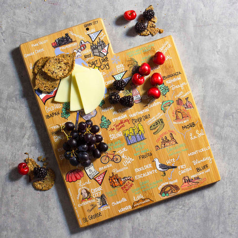 The Utah Cutting Board with Artwork by Fish Kiss™, from Totally Bamboo, is a state-shaped surface featuring vibrant illustrations of landmarks and city names along a delightful food trail. Beautifully arranged snacks, including cheese slices, cherries, grapes, blackberries, and crackers, grace the bamboo cutting board against its gray backdrop.