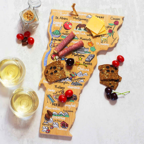 A Vermont-shaped cutting board by Totally Bamboo, featuring Fish Kiss™ artwork of towns, cheese, salami, crackers, and cherries carved from bamboo. Nearby, two glasses of white wine rest on a pristine white surface, perfectly accentuating the state-shaped board's natural charm.