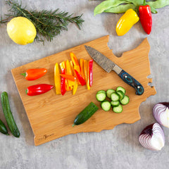 The Washington Cutting Board with Artwork by Fish Kiss™, crafted by Totally Bamboo, perfectly resembles the state of Washington. Sliced bell peppers and cucumbers add color to its surface, surrounded by whole vegetables such as onions and sprigs of rosemary. A knife lies on this vibrant bamboo board alongside bright peppers and a lemon, encapsulating the spirit of fresh culinary artistry.