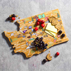 The Washington Cutting Board with Artwork by Fish Kiss™ from Totally Bamboo features a bamboo surface shaped like the state of Washington, adorned with names of its cities and landmarks. It is elegantly displayed with cherries, grapes, crackers, cheese, and blackberries on a stone surface.