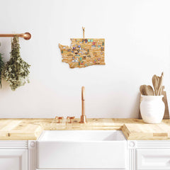 A modern kitchen features a sleek white farmhouse sink paired with a copper faucet, all set against a wooden countertop. Adding to the decor is the Totally Bamboo cutting board, adorned with unique artwork by Fish Kiss™, depicting a colorful map of Washington state. It hangs stylishly on the white wall above, amidst an array of herbs, utensils, and boards.