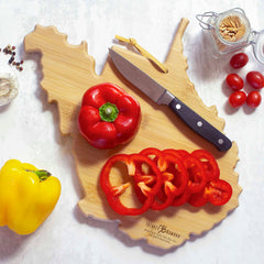 A West Virginia Cutting Board with Artwork by Fish Kiss™ from Totally Bamboo showcases sliced and whole red bell peppers alongside a knife. The clean white surface nearby holds a yellow bell pepper, cherry tomatoes, garlic, peppercorns, and a jar of toothpicks.