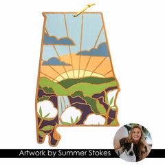 The Alabama-shaped wooden artwork, crafted by Summer Stokes and produced by Totally Bamboo, showcases a vibrant landscape complete with a sunset, clouds, green fields, and cotton plants. A circular inset highlights an image of a smiling woman with long hair alongside a dog. The text at the bottom reads "Alabama Cutting Board with Artwork by Summer Stokes.