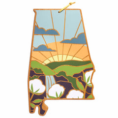 This Alabama Cutting Board with Artwork by Summer Stokes from Totally Bamboo is crafted from wood and shaped like the state of Alabama. It features an engraved design of a sunrise, clouds, fields, and cotton plants, and hangs elegantly by a yellow string.