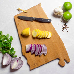 The Alabama Cutting Board by Totally Bamboo, featuring artwork by Summer Stokes, displays a partially sliced yellow squash and red onion with a nearby knife. On the white countertop around this artistic piece are lime, garlic, black pepper, cilantro, and additional onion.