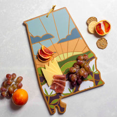 The Alabama Cutting Board with Artwork by Summer Stokes from Totally Bamboo is adorned with a vibrant landscape design reminiscent of Summer Stokes' artwork. This state-shaped cutting board beautifully presents slices of blood orange, cheddar cheese, and prosciutto, alongside grapes and crackers—a must-have for those who appreciate uniquely designed cutting boards.