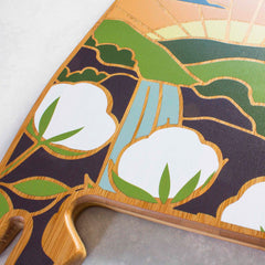 Introducing the Alabama Cutting Board by Totally Bamboo, adorned with a vibrant design by Summer Stokes. This visually striking piece showcases abstract green hills, a blue river, and white cotton plants set against dark backgrounds. Crafted from light wood and featuring a convenient grip, it seamlessly combines functionality with eye-catching artistry.